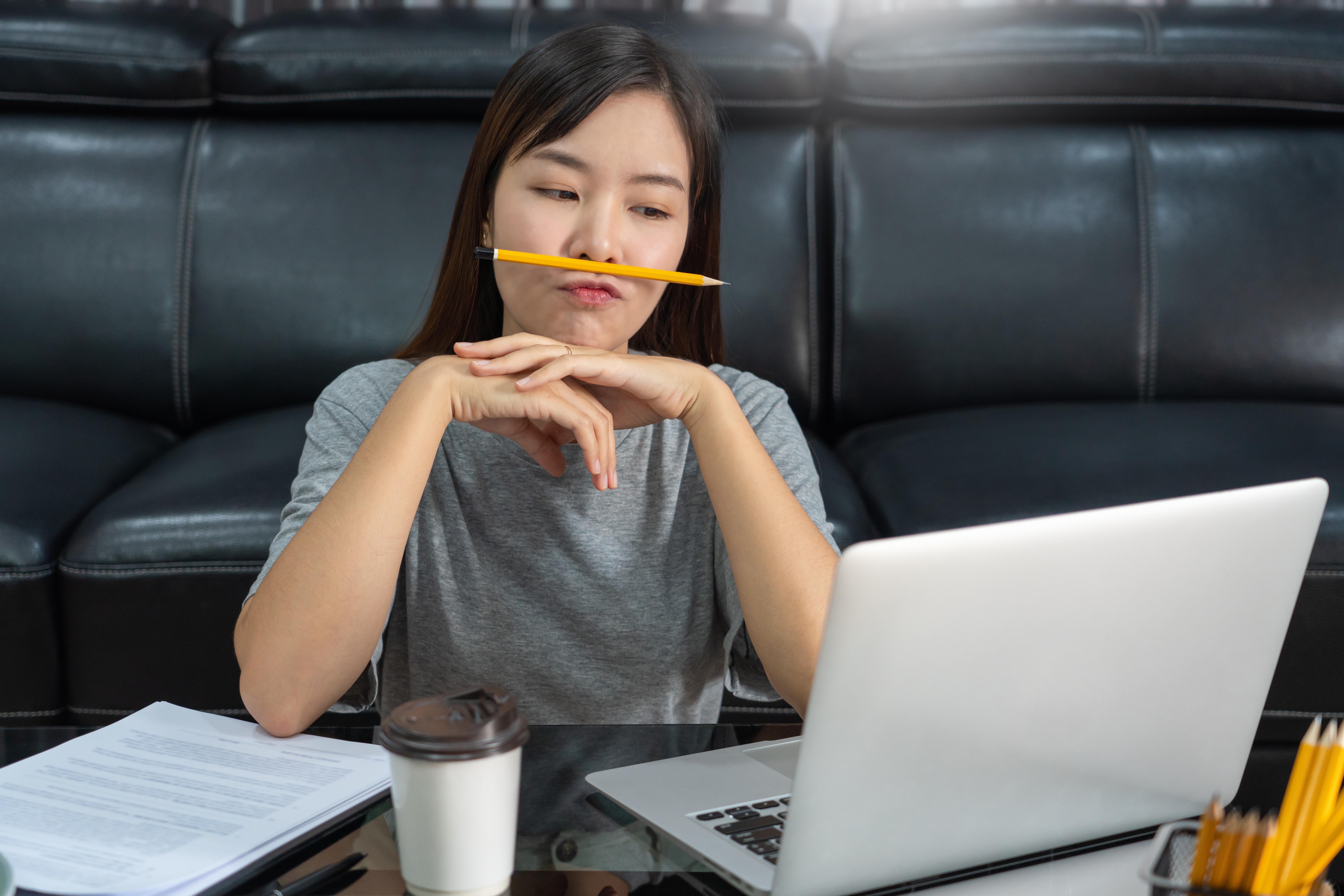 Young woman working on her laptop from a couch, pencil held thoughtfully at her mouth