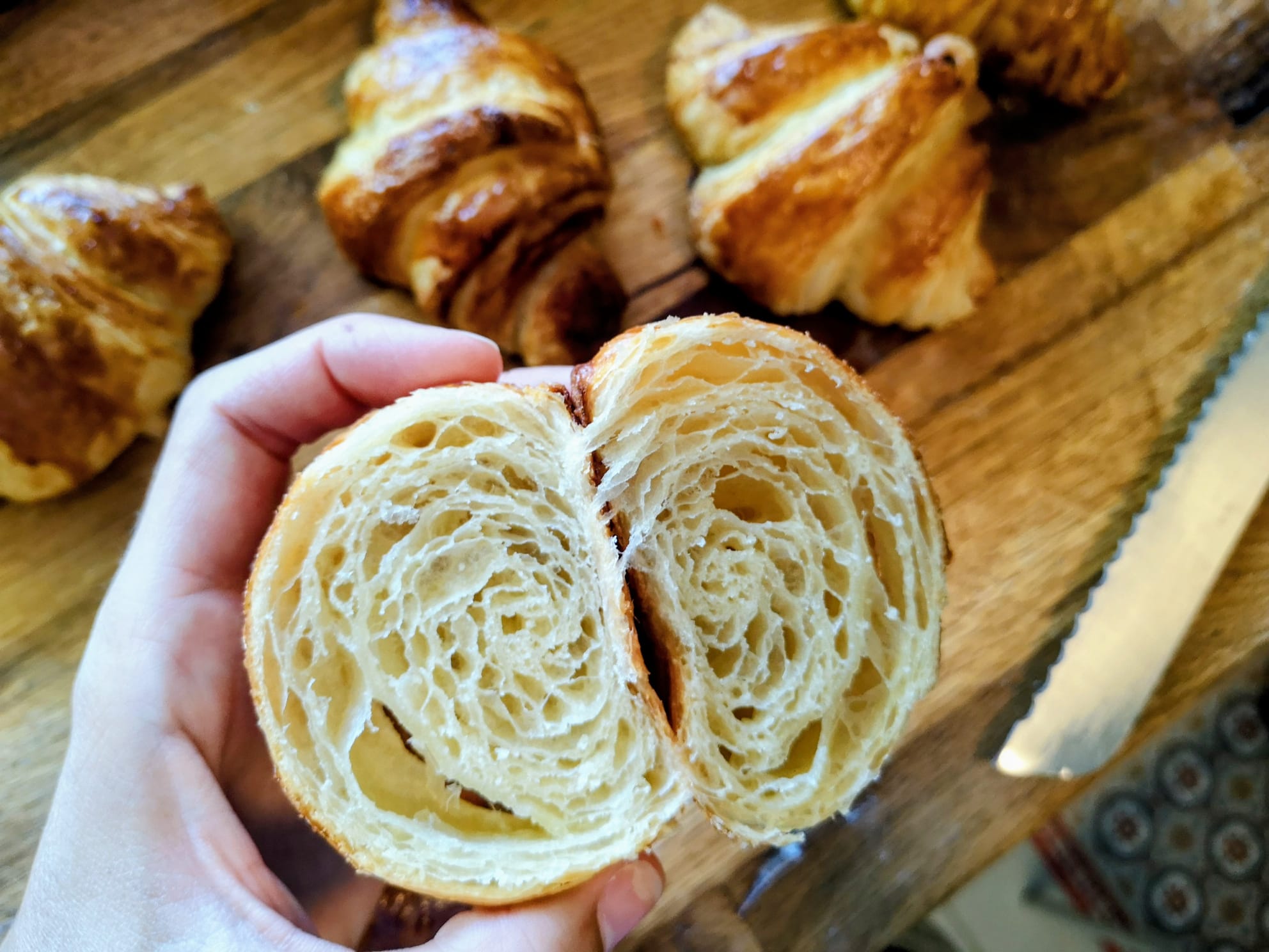 Hand-held halved croissant, cross-section showing the layered honeycomb interior, with whole croissants on a wooden board behind.